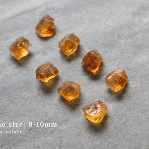 May include: Eight rough-cut citrine gemstones, each approximately 8-10mm in size, are scattered on a gray surface. The stones have a warm, amber color with variations in transparency and texture. The text "stone size: 8-10mm" is visible.