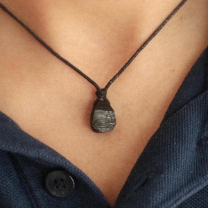 Puede incluir: Un collar con colgante negro con una piedra oscura de forma irregular suspendida de un cord&oacute;n negro. El collar se lleva sobre una camisa azul oscuro.