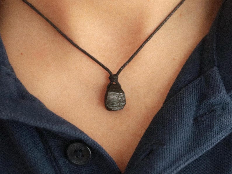 Puede incluir: Un collar con colgante negro con una piedra oscura de forma irregular suspendida de un cord&oacute;n negro. El collar se lleva sobre una camisa azul oscuro.
