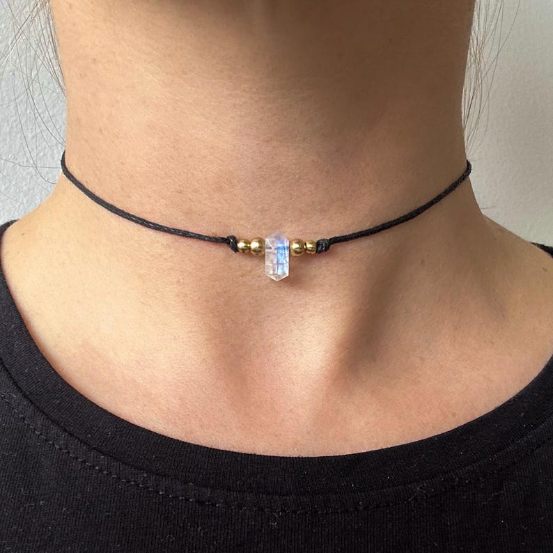 May include: A black choker necklace featuring a clear, rectangular crystal pendant with iridescent hues. The necklace has small gold-colored beads on either side of the crystal. The necklace is worn around the neck.