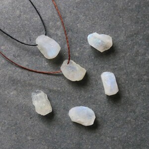Puede incluir: Una colecci&oacute;n de cuentas de piedra lunar en bruto e iridiscentes de varias formas y tama&ntilde;os. Dos collares con cordones negros y marrones est&aacute;n ensartados con piedras lunares. Las piedras est&aacute;n dispersas sobre una superficie gris oscuro, mostrando sus texturas naturales.