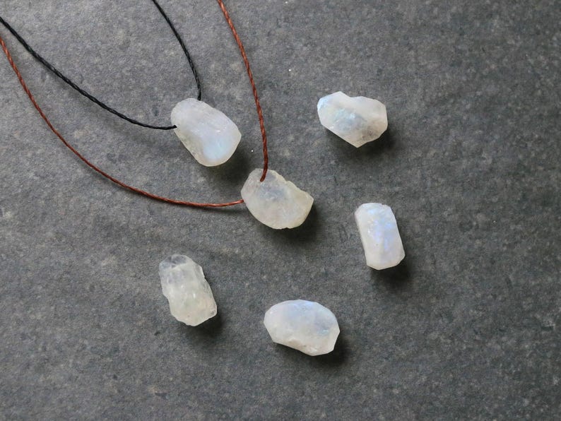 Puede incluir: Una colecci&oacute;n de cuentas de piedra lunar en bruto e iridiscentes de varias formas y tama&ntilde;os. Dos collares con cordones negros y marrones est&aacute;n ensartados con piedras lunares. Las piedras est&aacute;n dispersas sobre una superficie gris oscuro, mostrando sus texturas naturales.