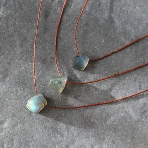 May include: Three necklaces featuring raw, iridescent blue-green labradorite stones. Each stone hangs from a thin, reddish-brown cord. The necklaces are presented on a textured, grey surface, highlighting the natural beauty of the gemstones.