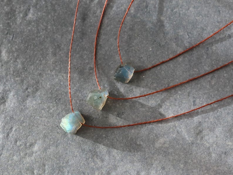 May include: Three necklaces featuring raw, iridescent blue-green labradorite stones. Each stone hangs from a thin, reddish-brown cord. The necklaces are presented on a textured, grey surface, highlighting the natural beauty of the gemstones.