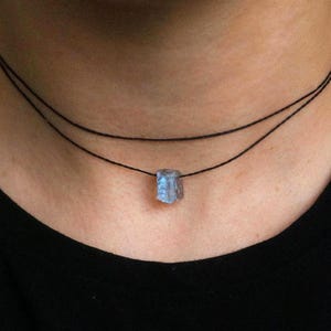 May include: A necklace with a small, pale blue gemstone pendant. The pendant hangs from two thin, black cords. The necklace is worn against a black top, which contrasts with the gemstone's colour and highlights its shape.