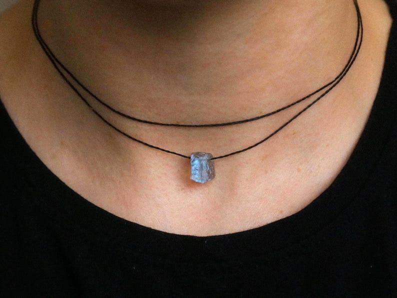 May include: A necklace with a small, pale blue gemstone pendant. The pendant hangs from two thin, black cords. The necklace is worn against a black top, which contrasts with the gemstone's colour and highlights its shape.