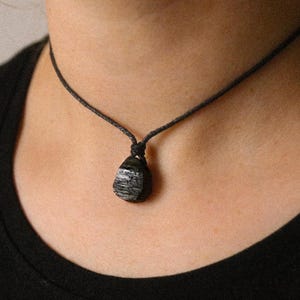 May include: A black cord necklace featuring a dark, textured pendant with gray and white streaks. The pendant is a rounded, teardrop shape. The necklace is worn against a black shirt.