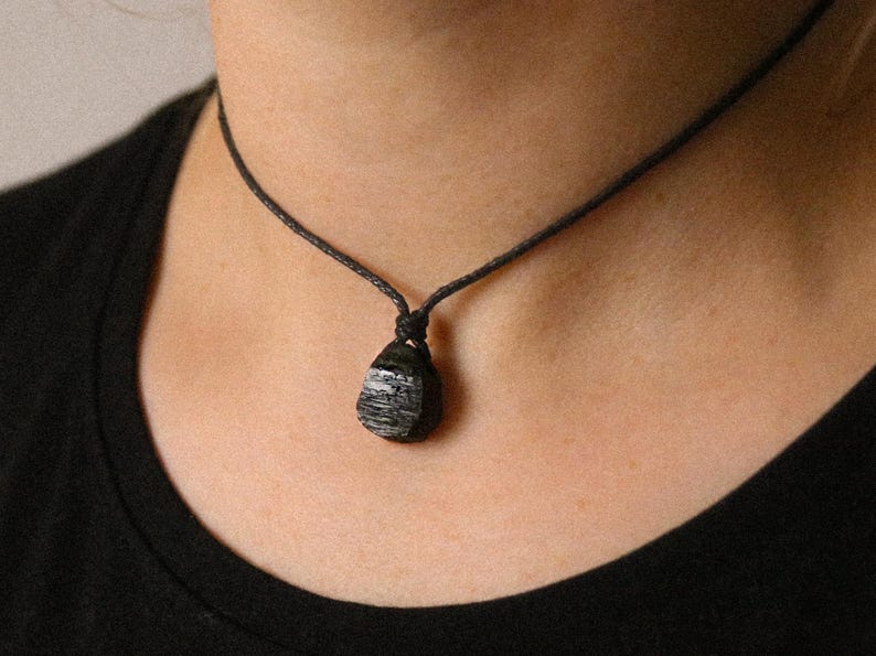 May include: A black cord necklace featuring a dark, textured pendant with gray and white streaks. The pendant is a rounded, teardrop shape. The necklace is worn against a black shirt.