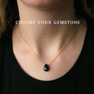 Puede incluir: Un collar de cadena dorada con un colgante de piedra preciosa negra en forma de lágrima. El collar se lleva sobre una parte superior negra. El texto "CHOOSE YOUR GEMSTONE" se muestra en blanco encima del collar.