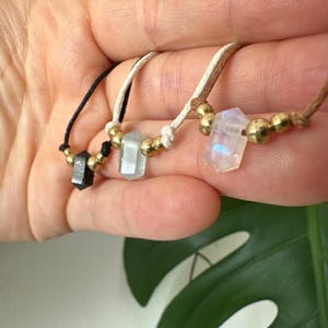May include: Three gemstone necklaces with gold beads. One necklace has a black crystal, another has a light blue crystal, and the third has a rainbow-hued crystal. The necklaces are strung on black, white, and brown cords.