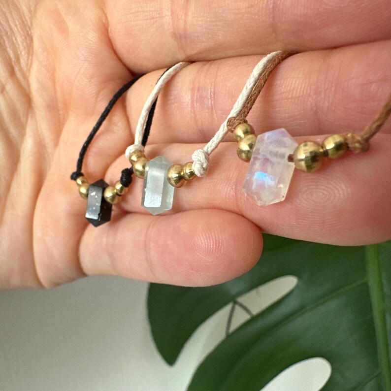 May include: Three gemstone necklaces with gold beads. One necklace has a black crystal, another has a light blue crystal, and the third has a rainbow-hued crystal. The necklaces are strung on black, white, and brown cords.