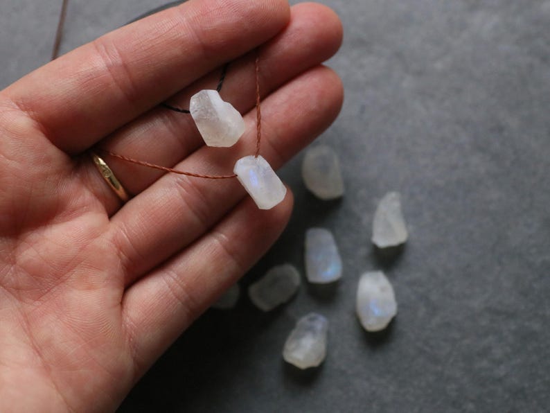 Puede incluir: Una mano sostiene un hilo de cuentas de piedra lunar blanca en bruto sobre un cord&oacute;n marr&oacute;n. Varios trozos sueltos de piedra lunar est&aacute;n esparcidos cerca. Las piedras tienen una textura rugosa y natural y un sutil brillo iridiscente.