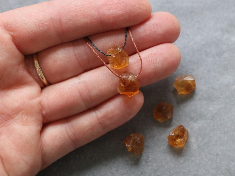 May include: A close-up of a necklace featuring two raw, orange-brown citrine gemstones strung on a thin cord. Several loose citrine stones are scattered nearby. The gemstones have a rough, natural texture and varying shapes.