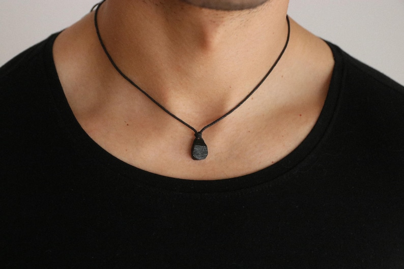 Puede incluir: Un collar con colgante negro con una piedra oscura y texturizada. El colgante cuelga de un fino cord&oacute;n negro. El collar se lleva sobre una camiseta negra.