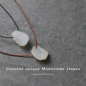 Puede incluir: Dos colgantes de piedra lunar blanca en bruto sobre cordones marrones y negros. Las piedras tienen forma irregular y un aspecto ligeramente transl&uacute;cido. El texto "Genuine unique Moonstone stones" y "crystalsofart" es visible.