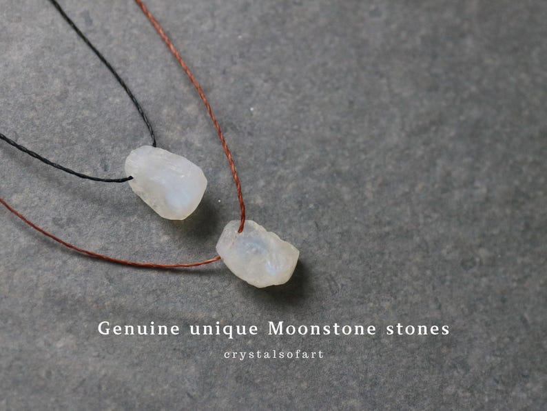 Puede incluir: Dos colgantes de piedra lunar blanca en bruto sobre cordones marrones y negros. Las piedras tienen forma irregular y un aspecto ligeramente transl&uacute;cido. El texto "Genuine unique Moonstone stones" y "crystalsofart" es visible.