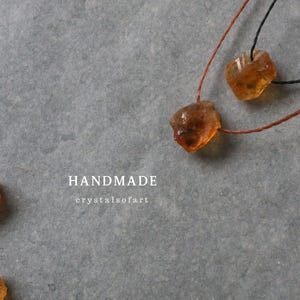 May include: Handmade citrine crystal necklaces with raw, amber-colored stones. The necklaces are strung on brown and black cords, displayed on a gray surface. The word "HANDMADE" is visible in white text.