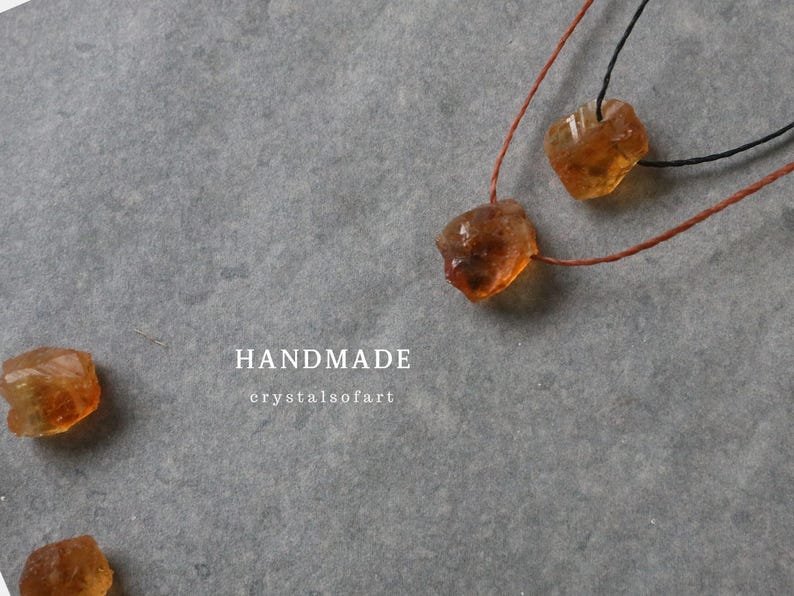 May include: Handmade citrine crystal necklaces with raw, amber-colored stones. The necklaces are strung on brown and black cords, displayed on a gray surface. The word "HANDMADE" is visible in white text.