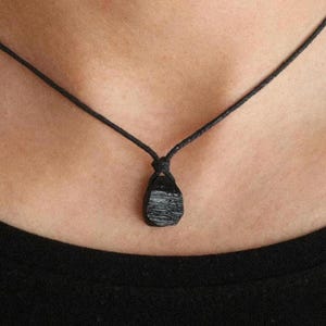 May include: A black tourmaline pendant necklace. The raw, textured gemstone is suspended from a black cord. The necklace is worn against a dark shirt.