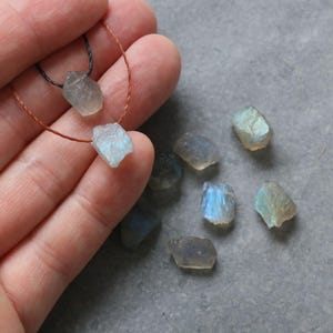 May include: Raw labradorite gemstone beads in various sizes and shapes. Two beads are strung on a thin cord, while others are loose. The stones display iridescent blue and green flashes. The background is a grey surface.