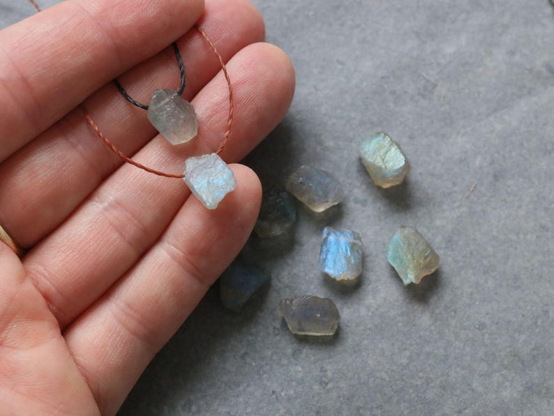 May include: Raw labradorite gemstone beads in various sizes and shapes. Two beads are strung on a thin cord, while others are loose. The stones display iridescent blue and green flashes. The background is a grey surface.