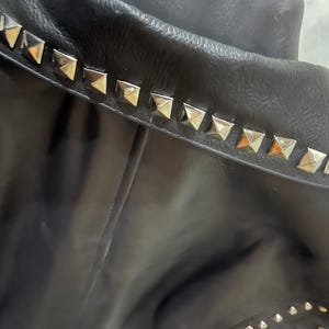 Puede incluir: Una chaqueta negra con un cuello ancho adornado con una hilera de tachuelas met&aacute;licas plateadas en forma de pir&aacute;mide. La prenda tiene una textura lisa e incluye tachuelas plateadas m&aacute;s peque&ntilde;as visibles en la parte inferior de la manga.