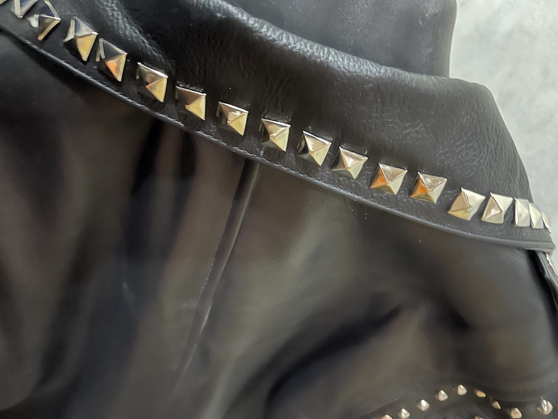 Puede incluir: Una chaqueta negra con un cuello ancho adornado con una hilera de tachuelas met&aacute;licas plateadas en forma de pir&aacute;mide. La prenda tiene una textura lisa e incluye tachuelas plateadas m&aacute;s peque&ntilde;as visibles en la parte inferior de la manga.