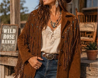 Puede incluir: Chaqueta de ante marrón con flecos y cuentas de colores. La chaqueta se lleva sobre un top de encaje crema y vaqueros azules. Un sombrero marrón y joyas completan el look western. Un letrero que dice "Wild Rose Ranch" es visible.
