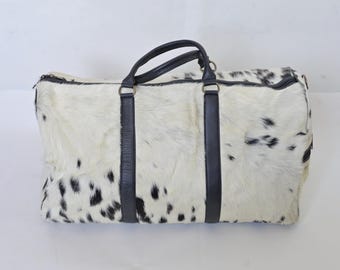 May include: A white and black cowhide duffel bag with black leather handles and straps.