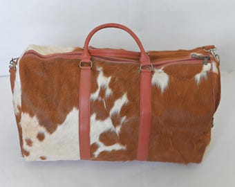 May include: A brown and white cowhide leather duffel bag with red leather handles and straps.