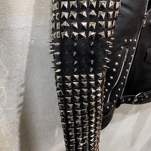 Puede incluir: Una manga de chaqueta negra densamente adornada con filas de tachuelas piramidales de metal plateado y p&uacute;as c&oacute;nicas afiladas. La prenda cuenta con un cierre de cremallera y un cintur&oacute;n con hebilla en la cintura sobre una superficie negra.