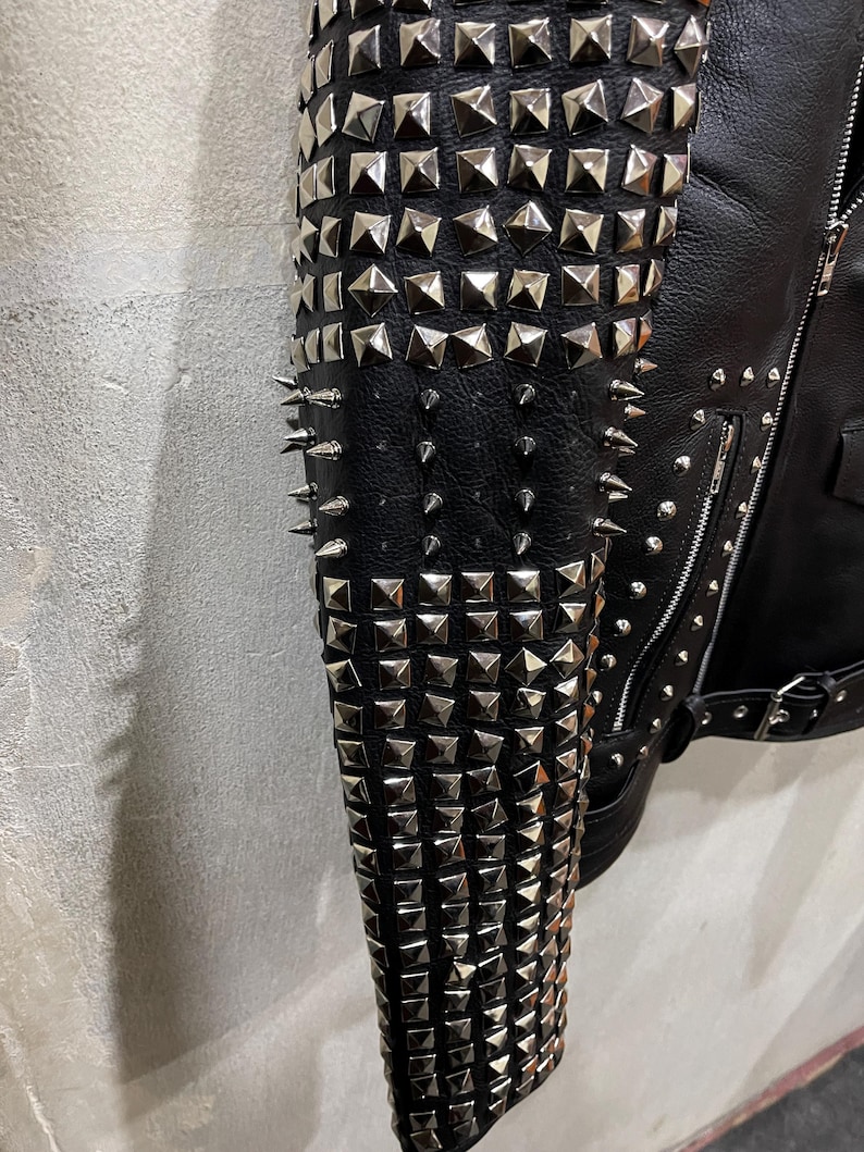 Puede incluir: Una manga de chaqueta negra densamente adornada con filas de tachuelas piramidales de metal plateado y p&uacute;as c&oacute;nicas afiladas. La prenda cuenta con un cierre de cremallera y un cintur&oacute;n con hebilla en la cintura sobre una superficie negra.
