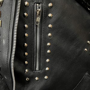 Puede incluir: Una chaqueta de cuero negro con un bolsillo vertical con cremallera en el pecho. La chaqueta est&aacute; decorada con numerosos remaches de metal plateado dispuestos en filas alrededor del bolsillo y a lo largo de los bordes. Se ve un tirador de cremallera YKK.
