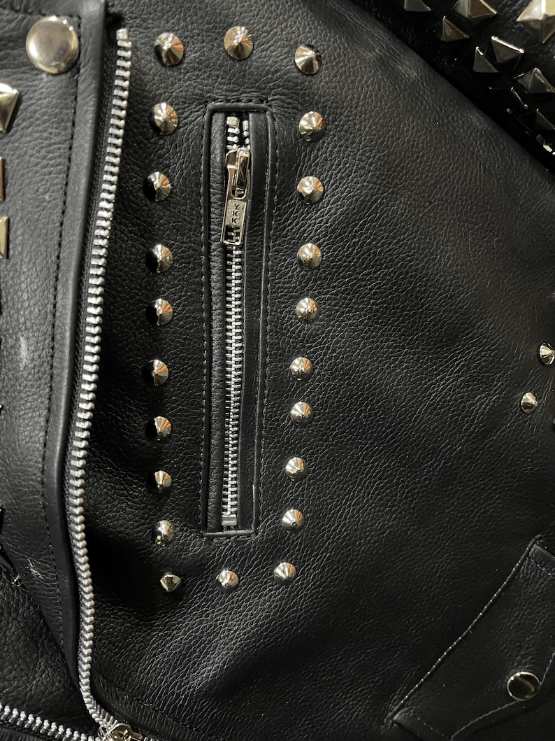 Puede incluir: Una chaqueta de cuero negro con un bolsillo vertical con cremallera en el pecho. La chaqueta est&aacute; decorada con numerosos remaches de metal plateado dispuestos en filas alrededor del bolsillo y a lo largo de los bordes. Se ve un tirador de cremallera YKK.