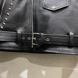 Puede incluir: Una chaqueta de cuero negro con tachuelas met&aacute;licas plateadas en los paneles frontales, una cremallera central plateada, un bolsillo con solapa y bot&oacute;n a presi&oacute;n, y un cintur&oacute;n de cuero ajustable con hebilla met&aacute;lica plateada.