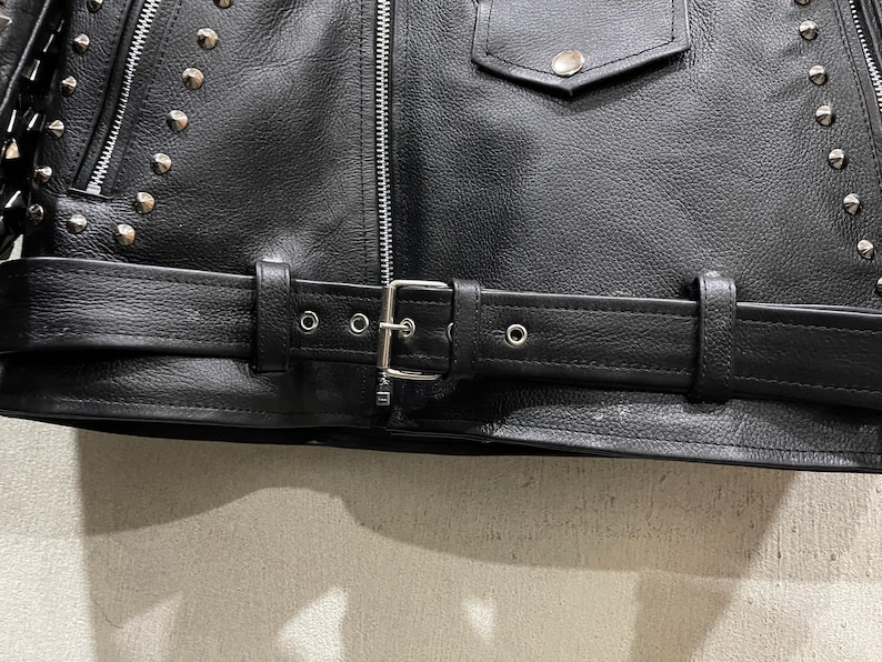Puede incluir: Una chaqueta de cuero negro con tachuelas met&aacute;licas plateadas en los paneles frontales, una cremallera central plateada, un bolsillo con solapa y bot&oacute;n a presi&oacute;n, y un cintur&oacute;n de cuero ajustable con hebilla met&aacute;lica plateada.