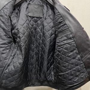 Puede incluir: Una chaqueta de cuero negro con forro interior acolchado y tachuelas piramidales de metal plateado que decoran los pu&ntilde;os. La chaqueta se muestra en una percha negra, revelando la construcci&oacute;n interior y el exterior de cuero texturizado.