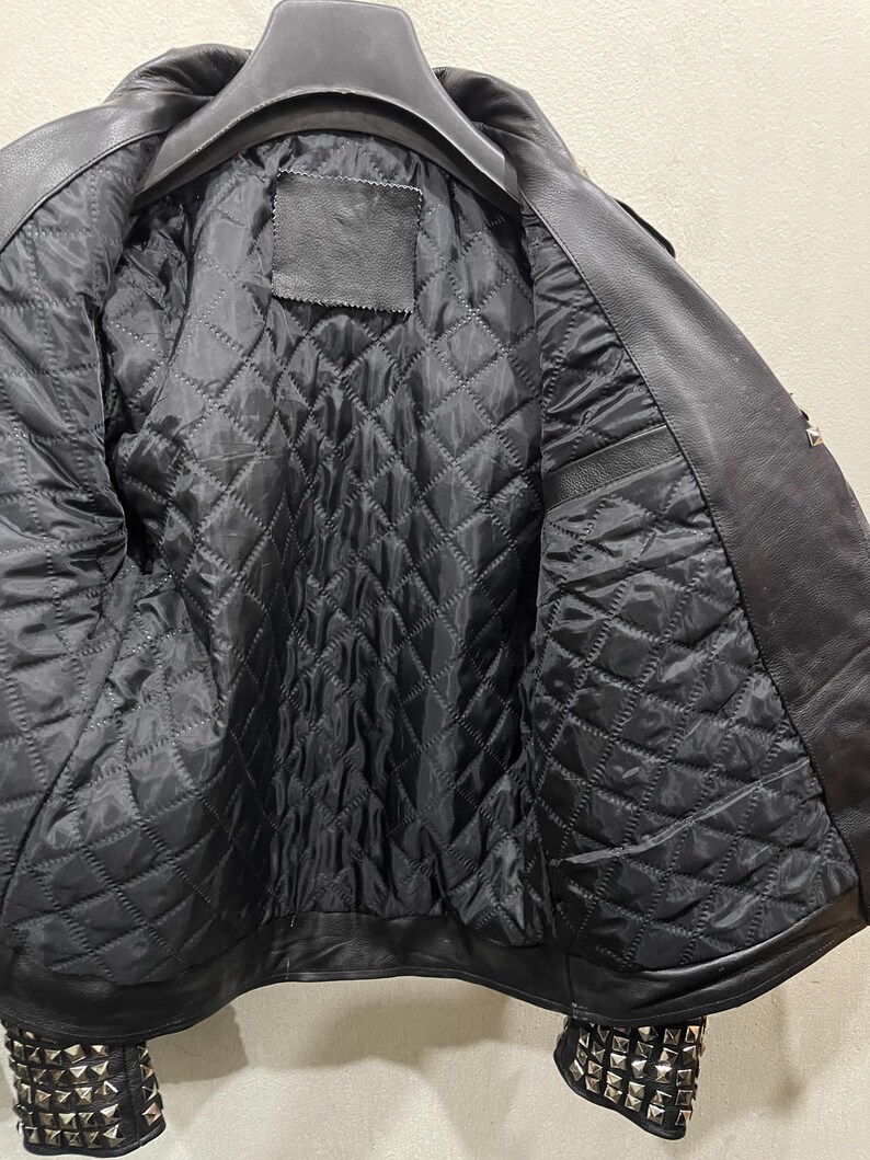 Puede incluir: Una chaqueta de cuero negro con forro interior acolchado y tachuelas piramidales de metal plateado que decoran los pu&ntilde;os. La chaqueta se muestra en una percha negra, revelando la construcci&oacute;n interior y el exterior de cuero texturizado.