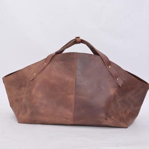 May include: A large, open-top tote bag made of brown leather. The bag has two handles and is constructed with visible stitching and rivets. The leather has a distressed appearance, with variations in color and texture.