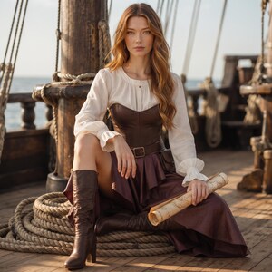 K&ouml;nnte beinhalten: Eine cremefarbene Langarmbluse mit ger&uuml;schten &Auml;rmelabschl&uuml;ssen, kombiniert mit einem dunkelbraunen Lederkorsett, einem passenden braunen Rock und kniehohen braunen Lederstiefeln. Das Outfit umfasst einen braunen Lederg&uuml;rtel und eine aufgerollte Karte.