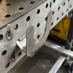 May include: A metal hook welded to a metal surface. The hook is shaped like a U and is designed to hold objects.