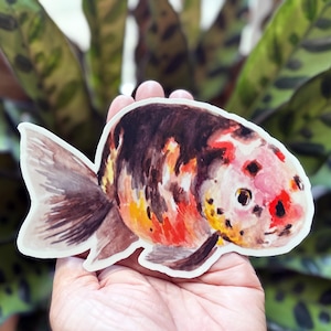 May include: A colorful sticker of a goldfish with a watercolor effect. The fish has a mix of red, orange, brown, and white markings. The sticker is held in a hand against a background of green leaves.