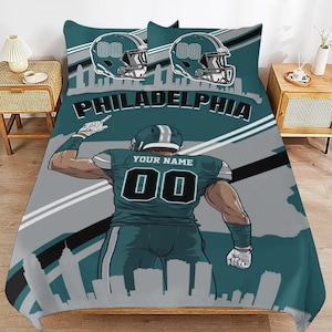 Könnte beinhalten: Türkis-graues Bettwäsche-Set zum Thema Football mit einer Spielerillustration, dem Text "PHILADELPHIA" und Footballhelmen. Das Set enthält einen Bettbezug und zwei Kissenbezüge. Das Trikot des Spielers hat die Nummer "00" und Platz für einen Namen.
