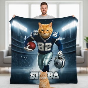 May include: A blue and white football-themed blanket featuring a cat wearing a football uniform with the number 82. The blanket has a stadium background and the word "SIBA" at the bottom. An orange cat is sitting in front of the blanket.
