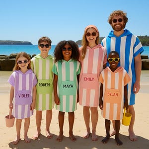 Könnte beinhalten: Eine Gruppe von Menschen, die passende gestreifte Kapuzen-Badetücher in verschiedenen Farben tragen, darunter Lila, Grün, Rosa und Orange. Jedes Handtuch hat eine Tasche mit einem aufgedruckten Namen. Sie stehen an einem Sandstrand.