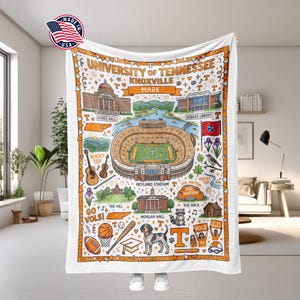 May include: A white University of Tennessee Knoxville blanket with an orange border. The blanket features a colorful illustration of campus landmarks, including Neyland Stadium, Ayres Hall, and Hodges Library. The words "University of Tennessee Knoxville" are at the top.