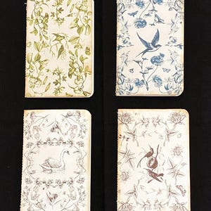 May include: Four small, blank, cream-colored notebooks with floral and bird designs. The notebooks have a vintage aesthetic and are bound with a brown string.
