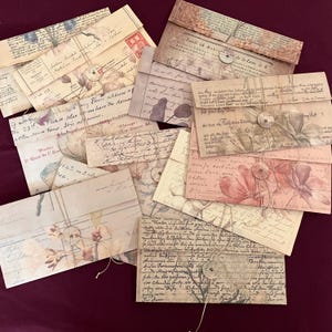 May include: A collection of vintage-style paper envelopes with floral and script designs. The envelopes are in various sizes and colors, including beige, cream, and pink. Each envelope is tied with twine and features a button closure, perfect for scrapbooking or letter writing.