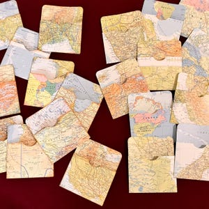 May include: A collection of square, vintage-style map cards scattered on a dark red surface. The cards feature detailed geographical illustrations in shades of beige, yellow, and blue, with a small pocket on each card.