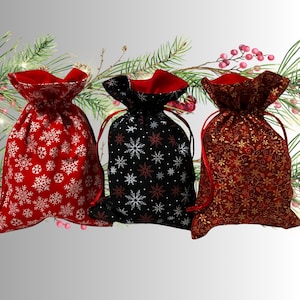 May include: Three Christmas gift bags in red, black, and burgundy. The red bag has white snowflakes, the black bag has white and red snowflakes, and the burgundy bag has gold floral patterns. Each bag has a red drawstring.